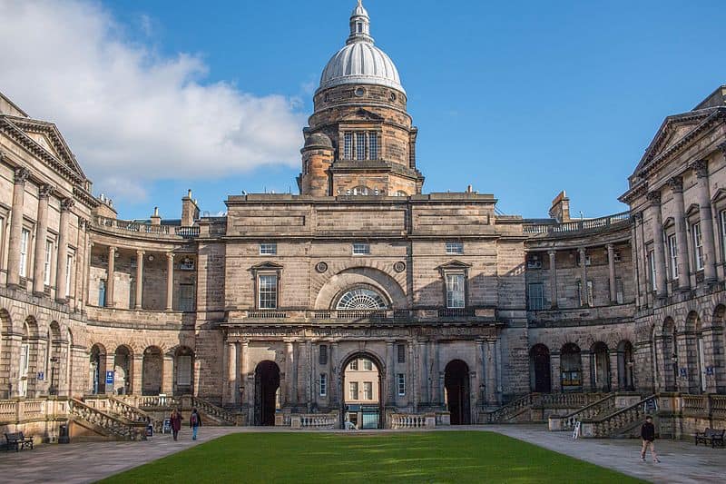 The Old College at the University of Edinburgh: a U-shaped grey stone two-storey academic building surrounds a large green lawn. The building's middle features a tower with a silver dome.