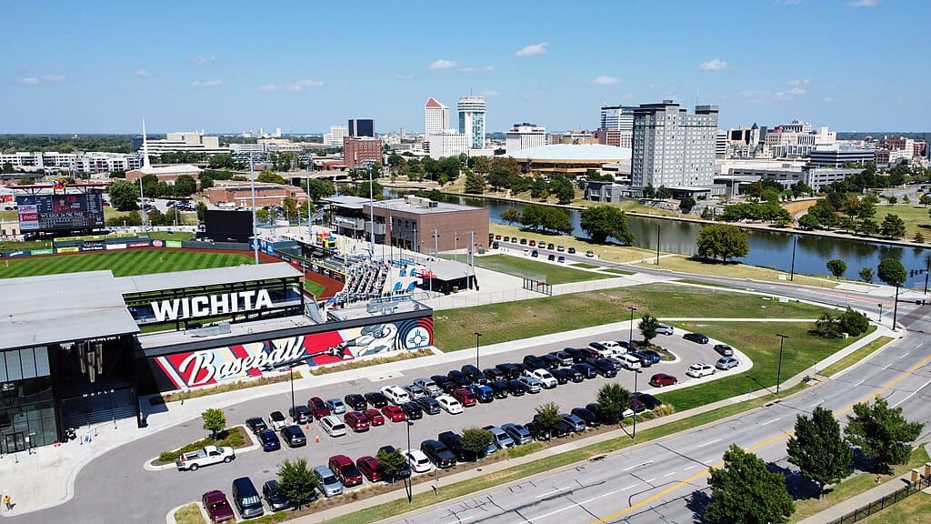 An aerial view of a large stadium with cars parked in front of it. Across a blue river is a mid-sized city skyline with office and apartment buildings.