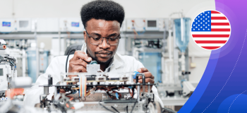 A young Black man works in an engineering lab environment, holding a soldering iron over a complex mechanical device.